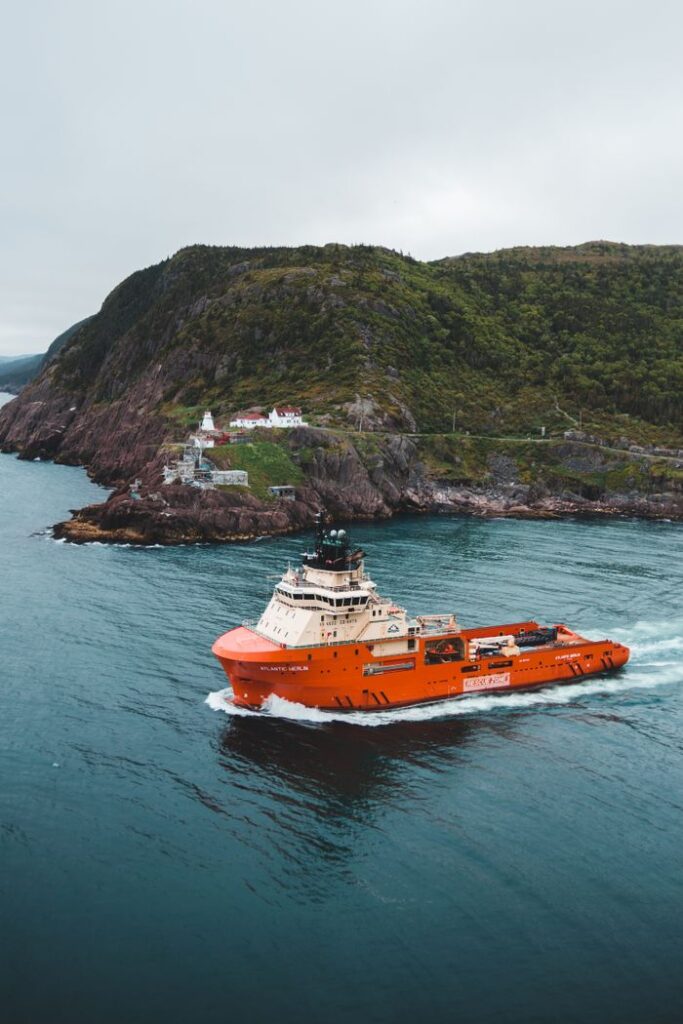 An orange cargo vessel navigates through calm sea waters near a rocky coastline with a lighthouse in view.